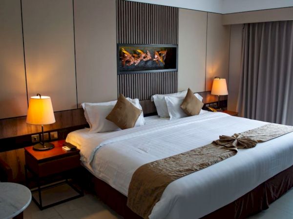 A well-lit hotel room with a king-sized bed, two bedside tables with lamps, a decorative fire display, and curtains covering a window.