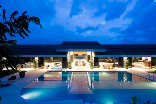 A luxurious resort or hotel illuminated at dusk, featuring a large swimming pool and an open, spacious lounge area.