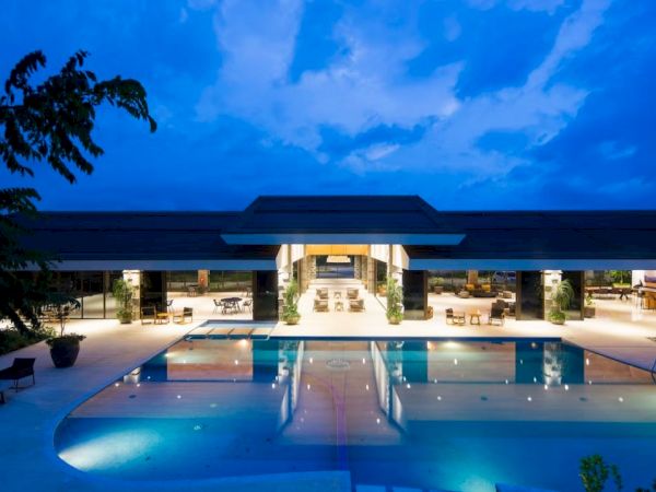 A luxurious resort or hotel illuminated at dusk, featuring a large swimming pool and an open, spacious lounge area.