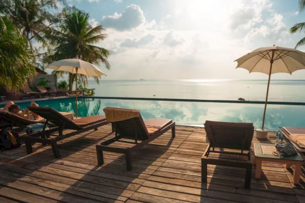 The image shows a tranquil poolside scene with lounge chairs, umbrellas, and a view of the ocean under a bright, partly cloudy sky.