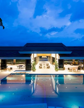 A luxurious outdoor pool area with a brightly lit modern building in the background and a clear evening sky overhead.