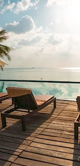 Lounge chairs and umbrellas on a wooden deck overlooking a serene infinity pool reflecting a partly cloudy sky, with a tropical beach in the background.