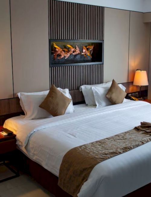 The image shows a cozy hotel room with a king-sized bed adorned with pillows and a decorative towel art, flanked by nightstands and lamps.