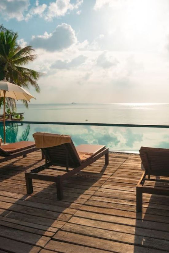 A serene poolside scene with lounge chairs, umbrellas, and a stunning view of the ocean, surrounded by lush tropical trees under a bright sky.