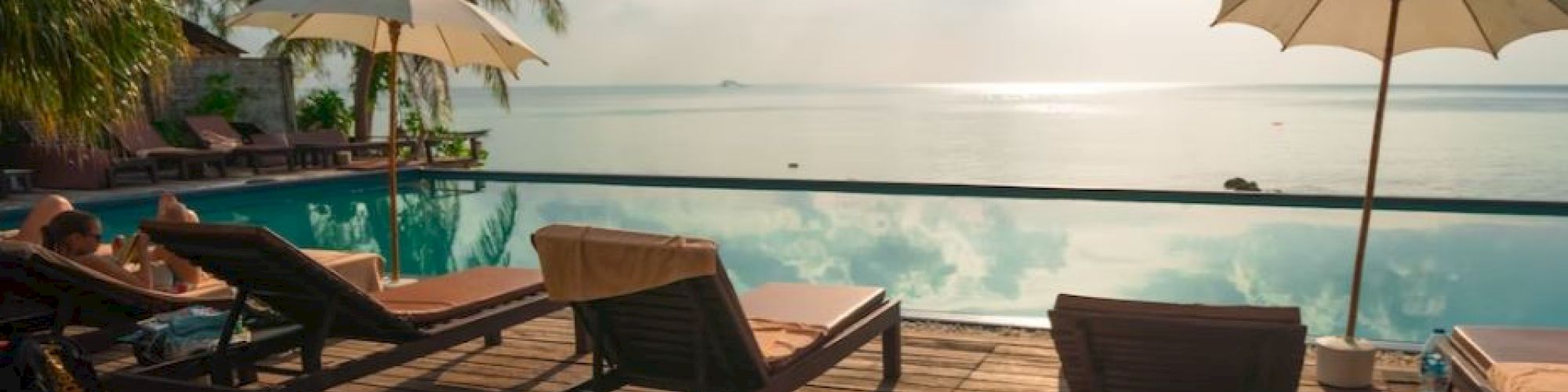 A serene poolside setup with sun loungers, umbrellas, and a view of the ocean under a partly cloudy sky.