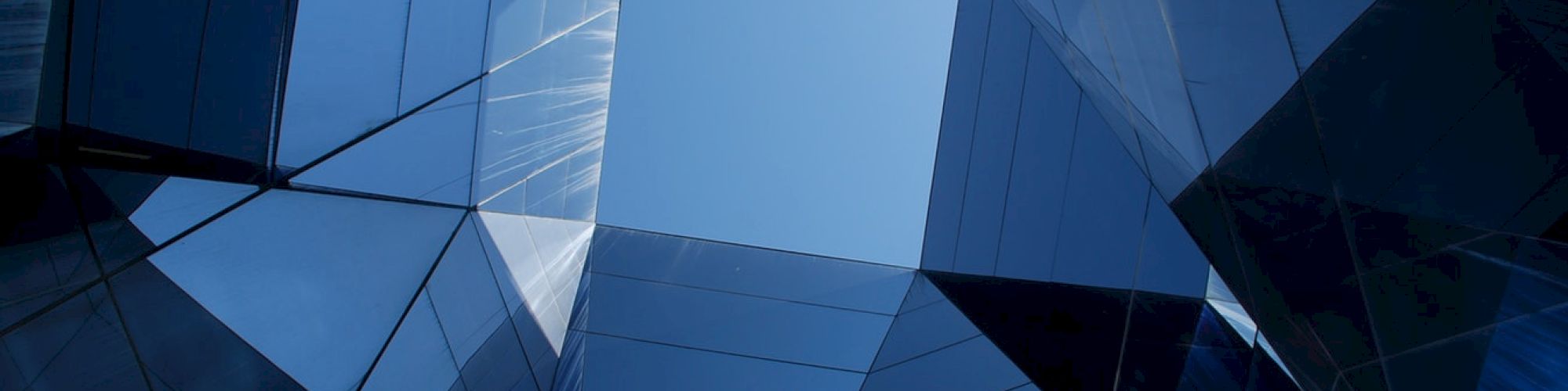 The image shows a view looking up through a geometric arrangement of glass buildings with reflections, creating a kaleidoscopic effect against the sky.