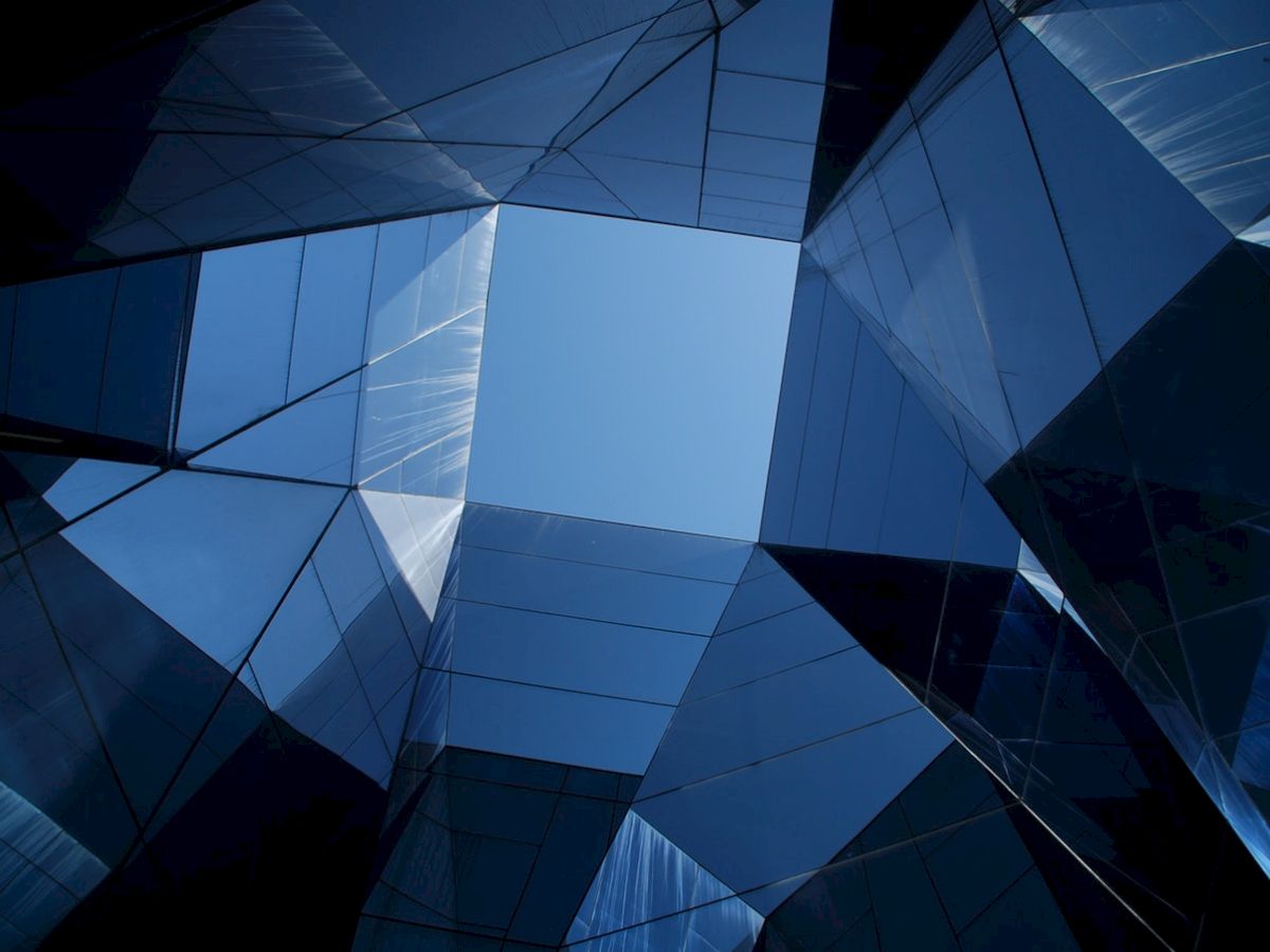 The image shows an upward view of tall, reflective glass buildings forming a geometric pattern with a clear blue sky in the center.