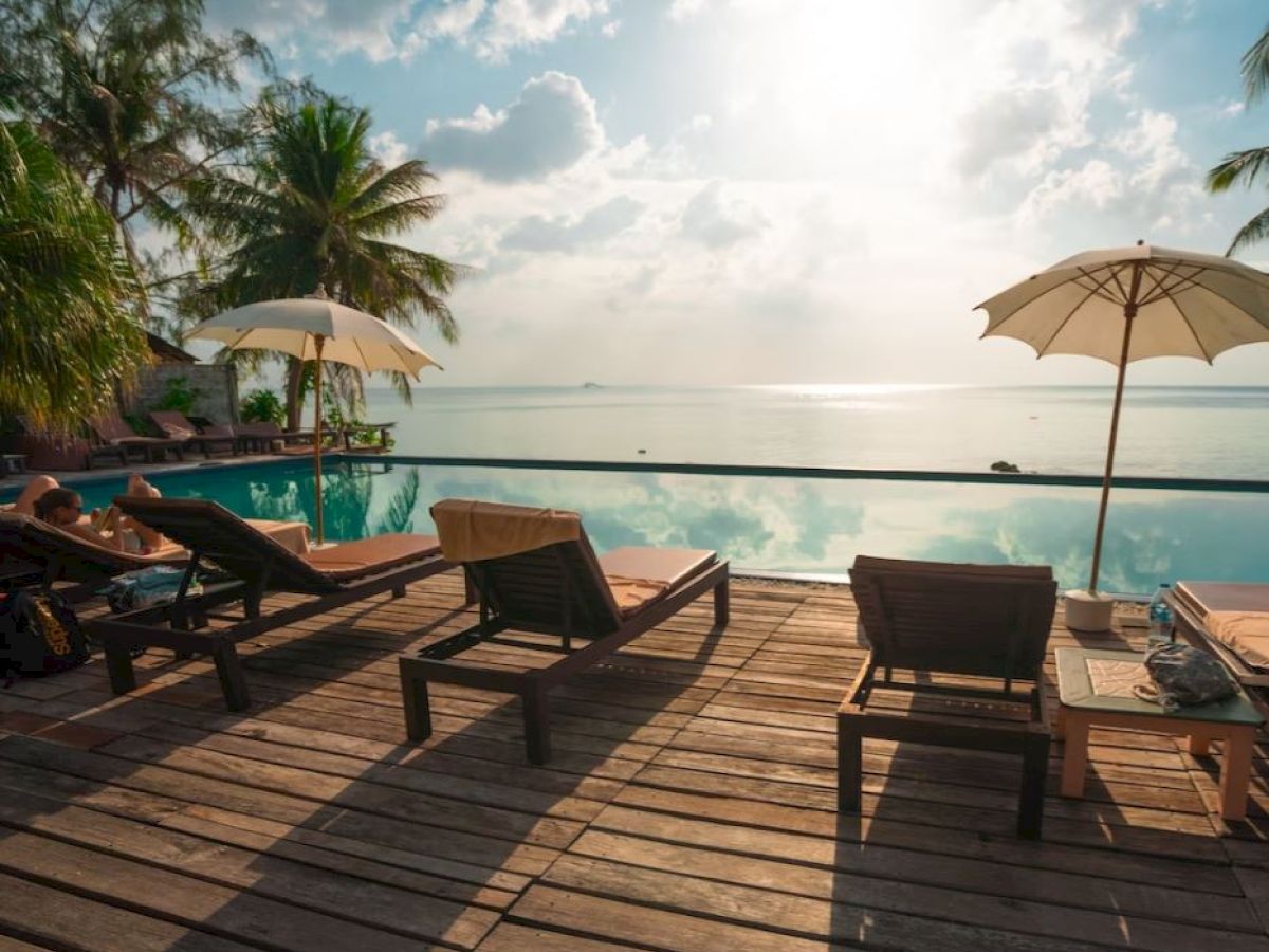 The image shows a serene poolside with wooden lounge chairs under umbrellas, facing a calm body of water during sunrise or sunset.