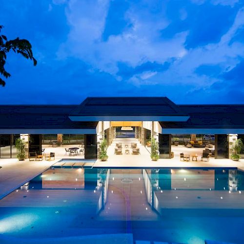 The image features a luxurious modern building with an illuminated swimming pool in the foreground under a vibrant evening sky.