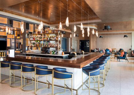 A modern bar with a stylish counter, blue bar stools, hanging lights, a variety of bottles on shelves, and a comfortable seating area.