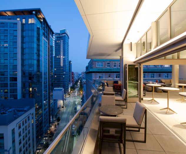 A modern cityscape at dusk with tall buildings, a street below, and an outdoor terrace featuring seating and tables in the foreground.