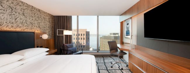 A modern hotel room features a large bed, a wall-mounted TV, a desk with a chair, and a window with a city view.