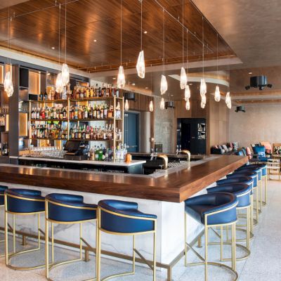 A modern bar area with blue upholstered stools, a well-stocked liquor shelf, hanging lights, and a cozy seating area in the background.