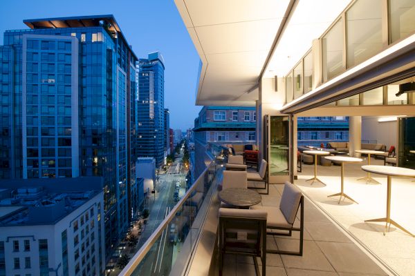 The image shows an outdoor balcony at dusk with modern furniture overlooking a cityscape with high-rise buildings and a street below.