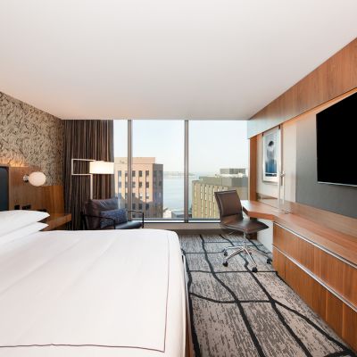 This is a modern hotel room featuring a large bed, desk, chair, wall-mounted TV, and a window with a city view. The room has stylish decor.