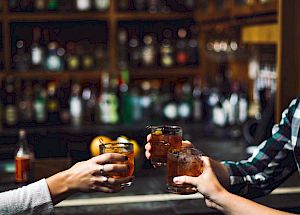 Three people in a bar clinking their glasses together, with shelves of bottles visible in the background.