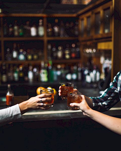 Three people in a bar clinking their glasses together, with shelves of bottles visible in the background.