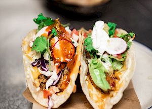 The image shows two tacos filled with various ingredients, including vegetables, sauce, and garnishes, placed on a plate atop brown paper.