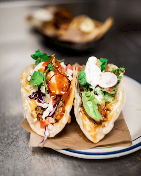 The image shows two tacos filled with various ingredients, including vegetables, sauce, and garnishes, placed on a plate atop brown paper.