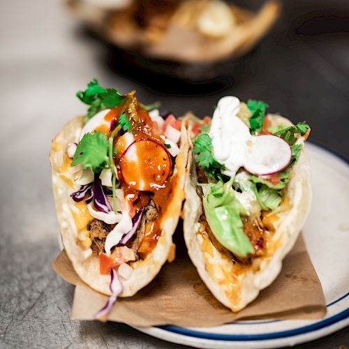 The image shows two tacos filled with various ingredients, including vegetables, sauce, and garnishes, placed on a plate atop brown paper.