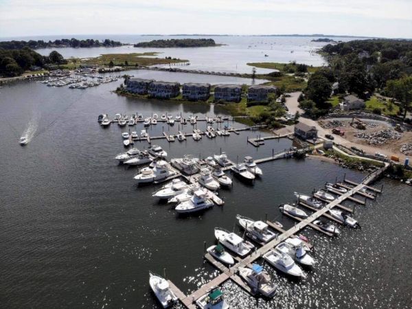 Aerial view of a marina with docks, multiple boats, and surrounding buildings, set against a coastal landscape with water and greenery.
