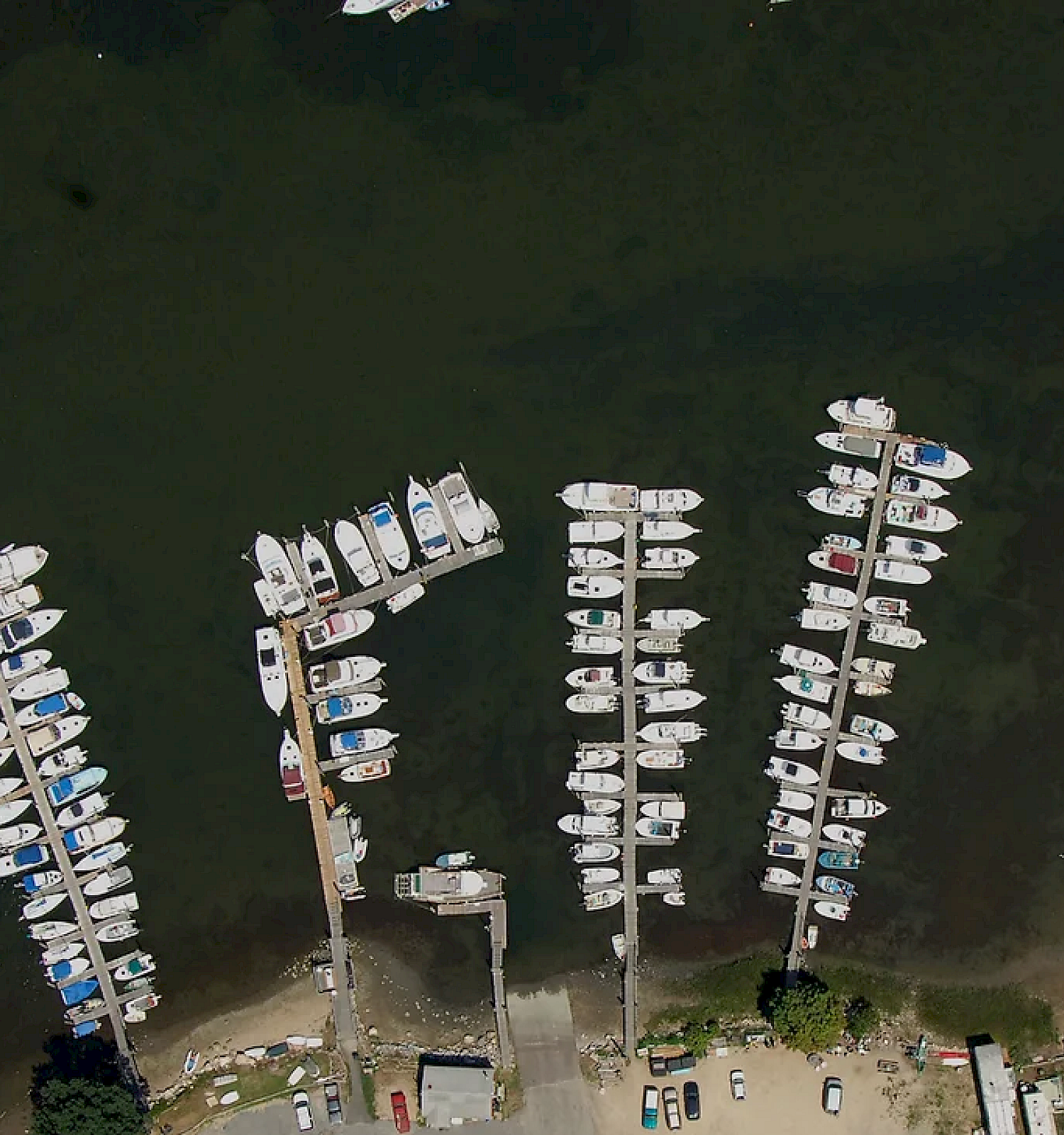 An aerial view of a marina with boats docked at piers, surrounded by parked cars and nearby buildings, on a body of water.