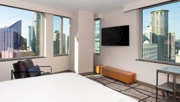 A modern hotel room with a large bed, TV, chair, and cityscape view from big windows. Ends the sentence.