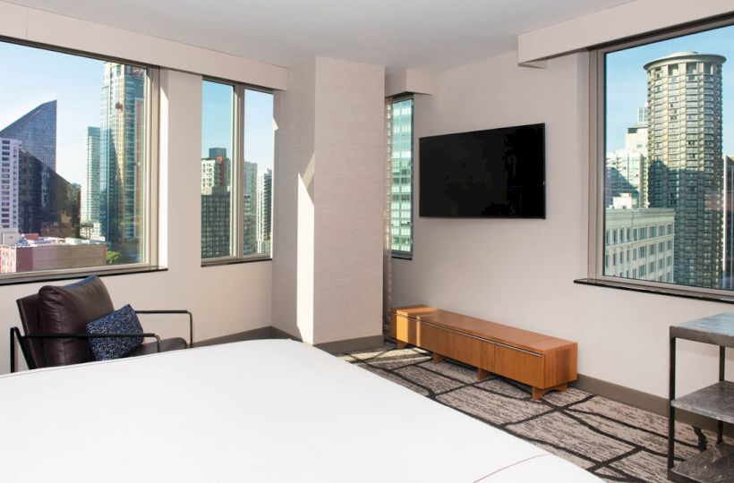 A modern hotel room with a large bed, TV, chair, and cityscape view from big windows. Ends the sentence.