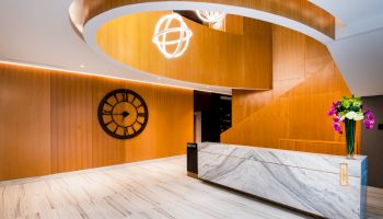 A modern, sleek lobby with a marble reception desk, wood accents, a spiral staircase, a large wall clock, and a flower vase on the desk.