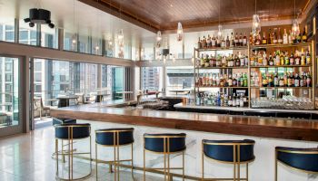 The image shows a modern bar with blue cushioned stools, a well-stocked liquor shelf, and large windows offering city views.