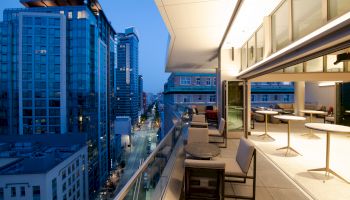 A modern cityscape view from a stylish, furnished balcony overlooking the street, with tall buildings and illuminated windows in the evening.