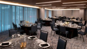 A banquet hall is set up with round tables covered with glasses, napkins, and cutlery, with black chairs surrounding each table.