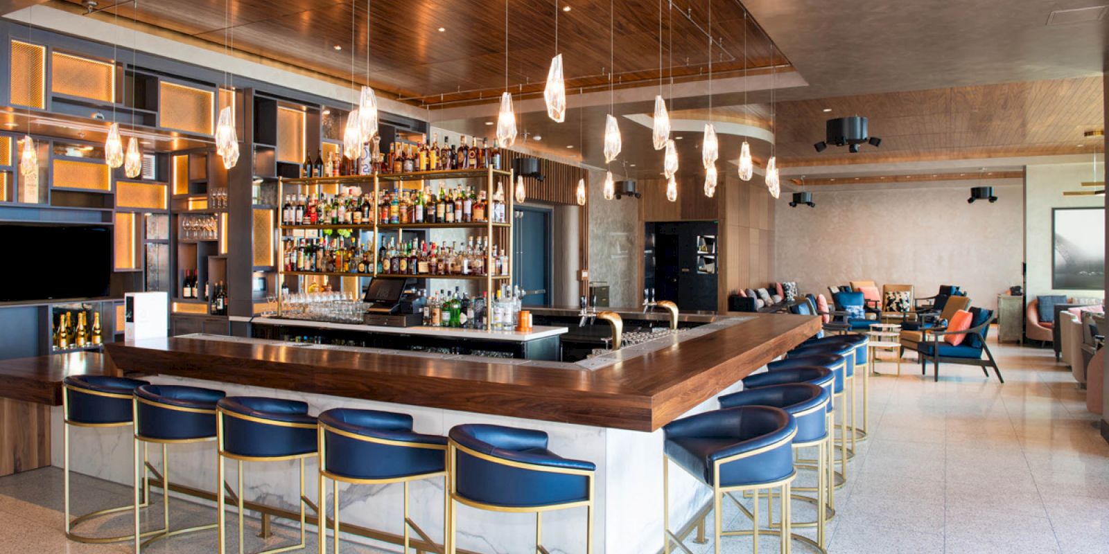 The image shows a modern bar with a wooden countertop surrounded by blue bar stools and a variety of bottles on shelves. Modern seating in the background.