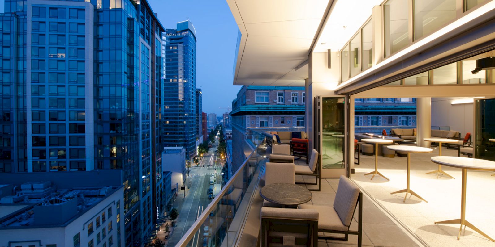 A modern balcony with tables and chairs overlooks a cityscape of tall buildings at twilight. The space is well-lit with an open seating area.