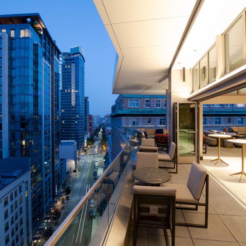 A modern balcony at dusk overlooks a cityscape with tall buildings; the area has seating and tables.