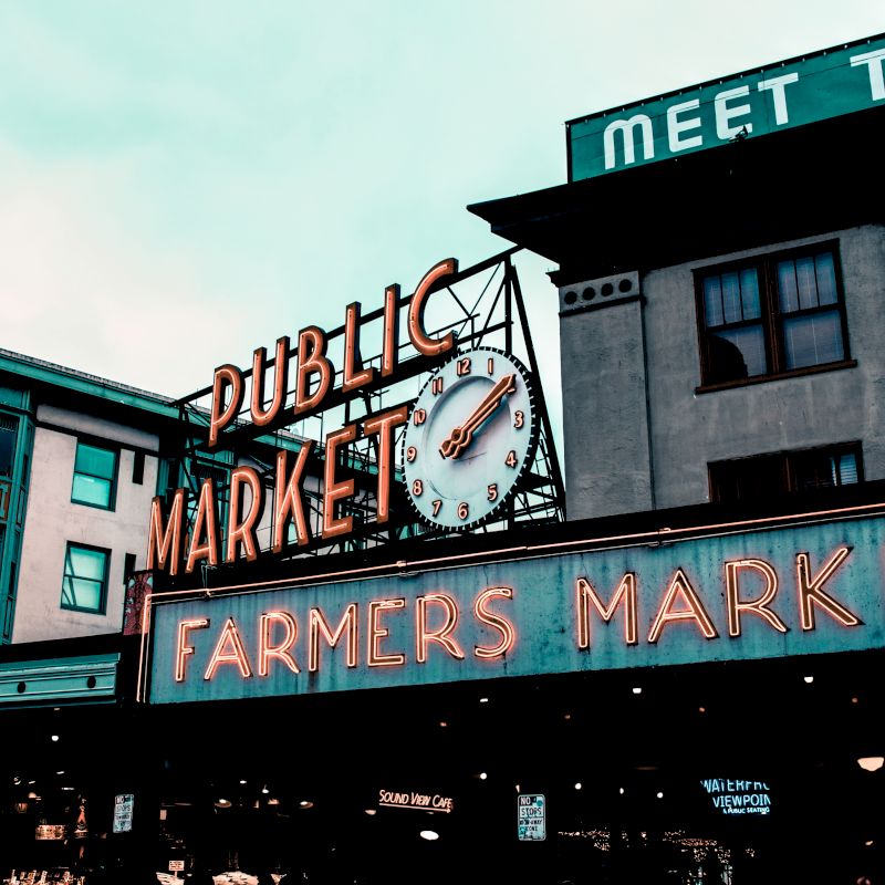 The image shows a vibrant sign for a public Farmers Market, featuring a clock, set against a backdrop of buildings, with one sign partially showing 