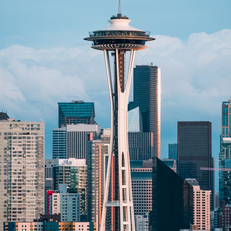 The image shows the Space Needle in Seattle, with various skyscrapers and buildings in the background, under a cloudy sky.