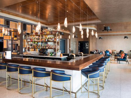 This image shows a modern bar with a well-stocked liquor shelf, elegant hanging lights, bar stools, and a seating area in the background.