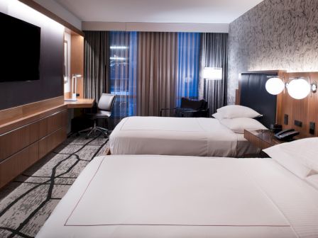 This image shows a modern hotel room with two neatly made beds, a flat-screen TV, a desk with a chair, and artistic decor.