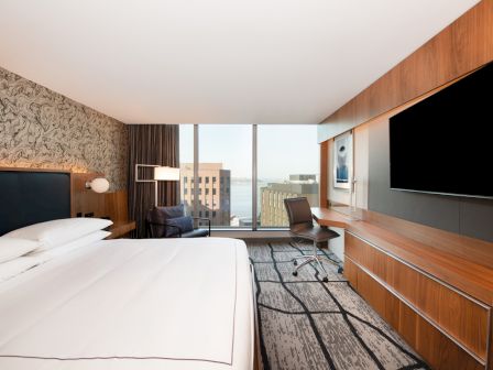 The image shows a modern hotel room with a large bed, a desk with a chair, a flat-screen TV, and a window view of city buildings and the water.