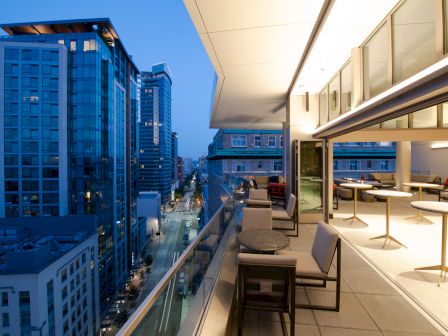 A modern cityscape at dusk showing high-rise buildings, a street below, and a rooftop terrace with tables and chairs, under a sleek overhang.