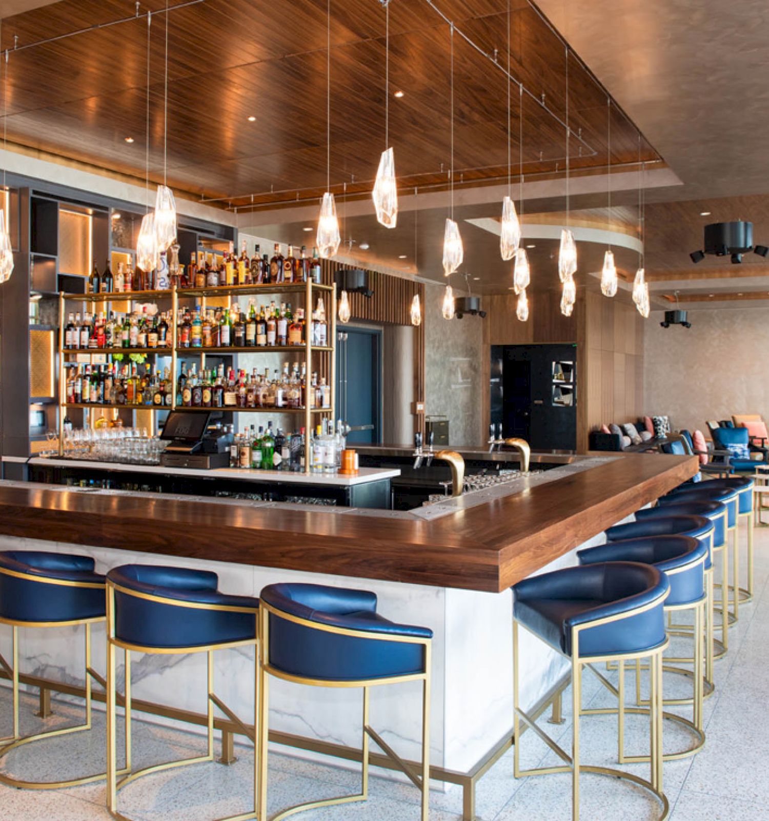 The image shows a modern bar with a wooden counter, blue barstools, and hanging lights. Shelves behind the bar are stocked with various bottles.