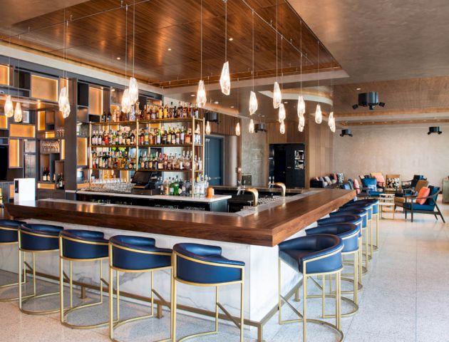 The image shows a modern bar with a wooden countertop, shelves stocked with bottles, pendant lights overhead, and blue bar stools arranged neatly.