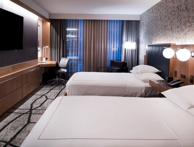 The image shows a modern hotel room with two neatly made beds, a wall-mounted TV, a work desk, and a window with curtains.