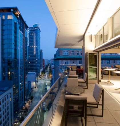 This image shows a modern cityscape at dusk featuring tall buildings and a contemporary balcony with seating and tables on the right side.