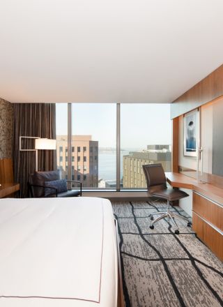 A modern hotel room features a large bed, a TV, a desk with a chair, and a view of city buildings and water through large windows.