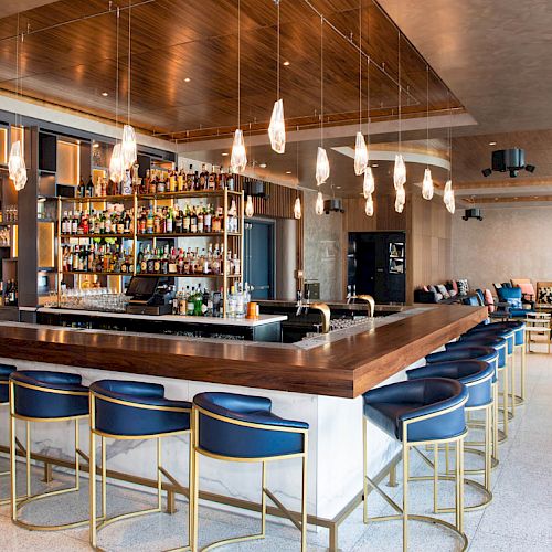 This image shows a modern bar with a wooden countertop, blue bar stools, and a display of various bottles behind the bar.