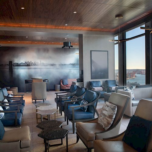 A modern lounge area with comfortable seating overlooks a scenic waterfront and city skyline through large windows on the right side.