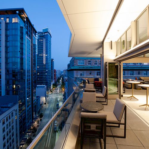 A modern cityscape at dusk shows high-rise buildings. The view includes an elegant terrace with tables, chairs, and ambient lighting.