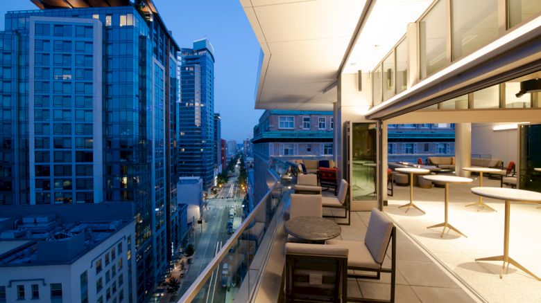 A rooftop balcony overlooking a cityscape at dusk, with outdoor seating and tables, and buildings lit up in the background.